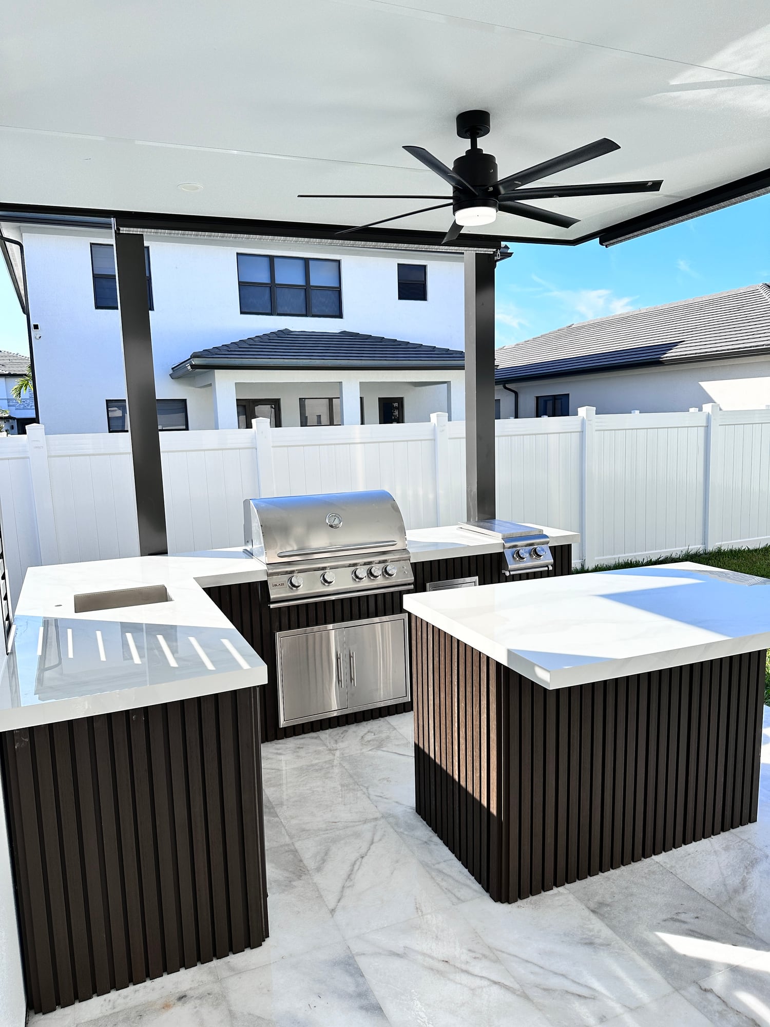 White outdoor kitchen countertop with a separate island and dark slatted cabinetry